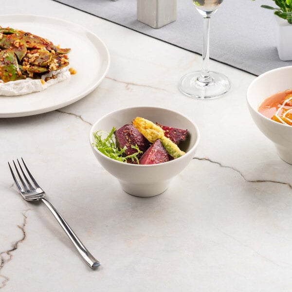 A small ivory porcelain bowl filled with roasted vegetables and greens is placed on a marble surface, surrounded by other dishes and a fork.