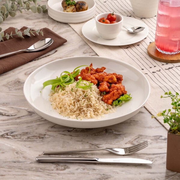A round, shallow ivory bowl filled with rice, fried chicken pieces, and garnished with green onions, placed on a marble table with cutlery and other dishes nearby.