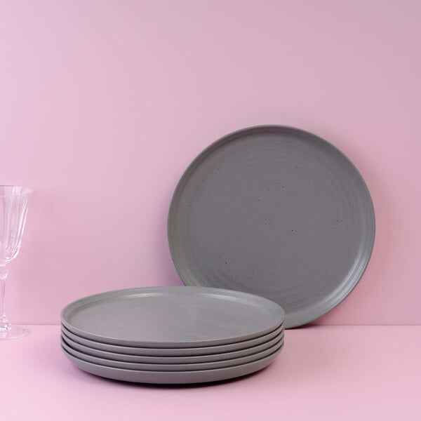 A stack of six round gray plates is displayed in front of a pink background, with one plate standing upright and a clear glass visible to the left.