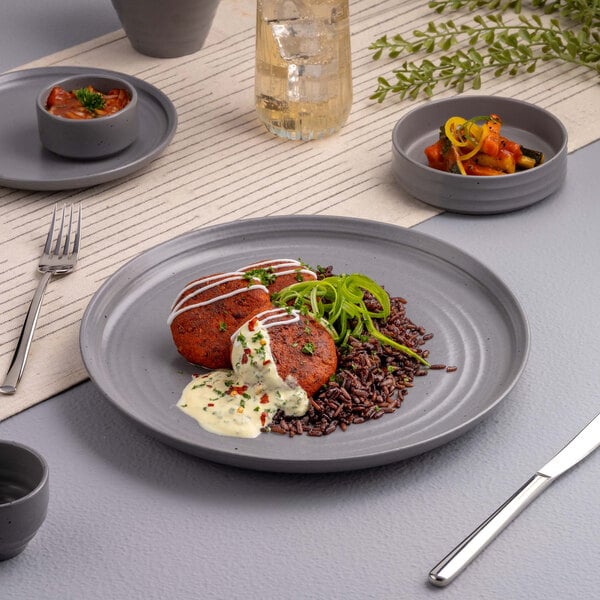 A gray round porcelain plate with two patties, a serving of dark rice, green garnish, and a creamy sauce, set on a modern table with matching gray dishware.