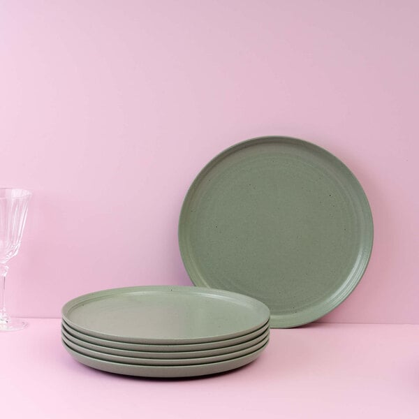 A stack of five green round plates with one plate standing upright behind them, set against a pink background with a clear glass on the left.