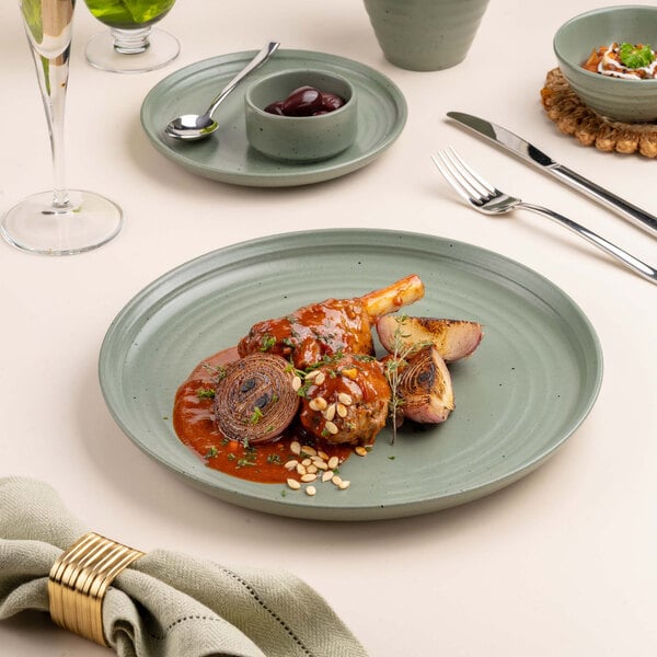 A green ceramic plate with a serving of braised meat in sauce, garnished with pine nuts and herbs, accompanied by roasted onions.