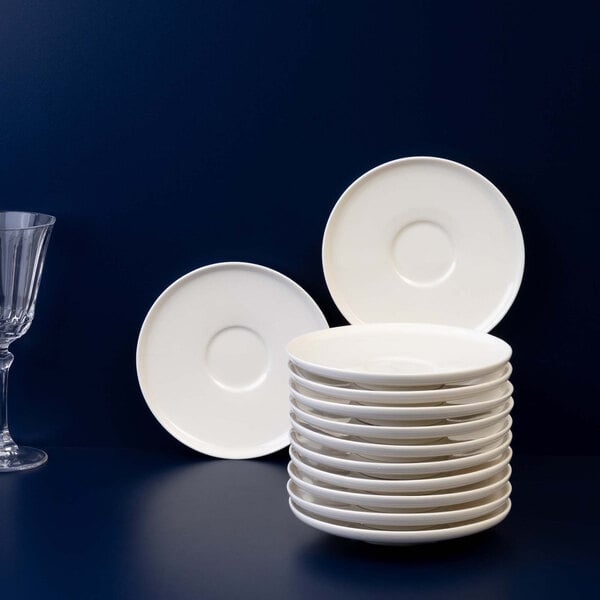 A stack of white round saucers is displayed on a dark surface, with two additional saucers standing upright and a clear glass on the left side.