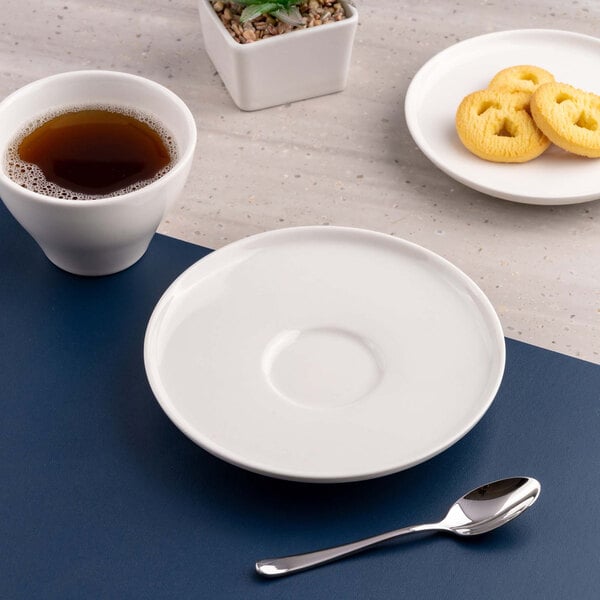 A white round saucer is placed on a blue surface next to a cup of coffee, a spoon, and a plate of cookies.