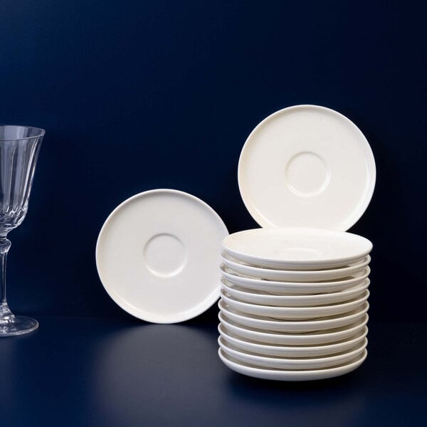 A stack of twelve round ivory saucers is displayed on a dark surface, with two additional saucers standing upright and a clear glass nearby.