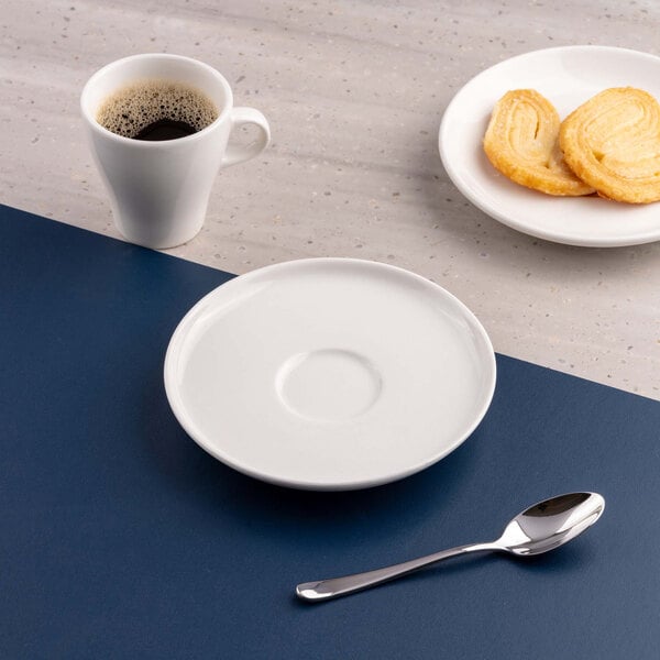 A white porcelain saucer is placed on a blue and gray surface, accompanied by a cup of coffee, a plate with cookies, and a spoon.