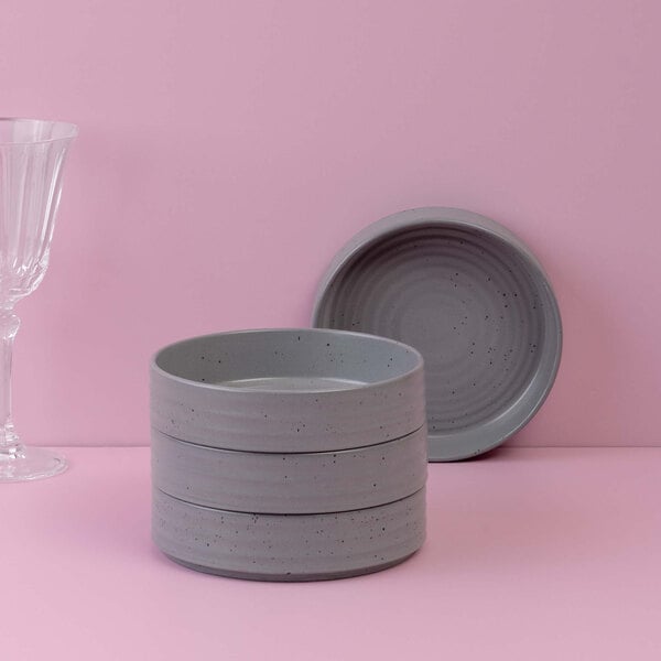 Four stacked round gray bowls with a speckled pattern, accompanied by a matching plate and a clear glass, all set against a pink background.