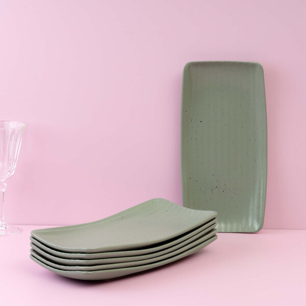 A stack of six rectangular green plates with a subtle speckled pattern is displayed on a pink surface, with one plate standing upright and a clear glass visible to the left.