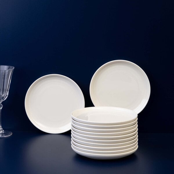 A stack of plain white round plates is displayed on a dark surface with two additional plates standing upright in the background and a clear glass on the left side.