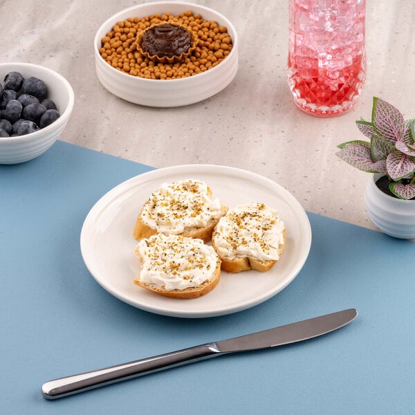 A white round plate with three pieces of bread topped with a creamy spread and herbs, placed on a blue placemat with a knife nearby; surrounding items include bowls of blueberries and snacks, a pink drink, and a small potted plant.