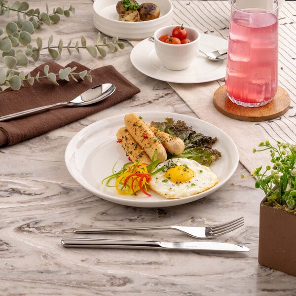 A round white plate with a fried egg, sausages, lettuce, and sliced peppers is set on a marble table with cutlery, a glass of pink beverage, and other small dishes nearby.