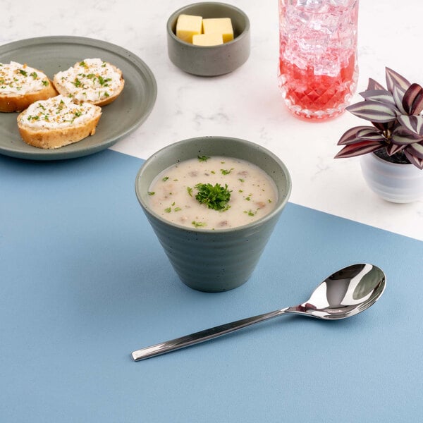 A green bowl filled with creamy soup garnished with herbs, placed on a blue surface next to a spoon, with bread, butter, a drink, and a small plant in the background.