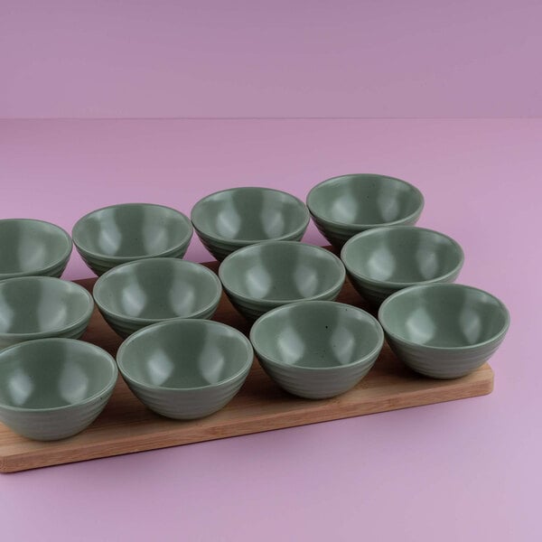 Twelve small green bowls are arranged on a wooden board against a pink background.