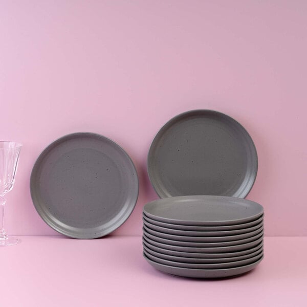 A stack of gray round plates is displayed in front of a pink background, with two plates standing upright and a glass partially visible on the left.