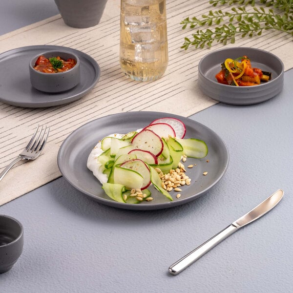 A gray round plate with a fresh salad of sliced radishes, cucumber ribbons, and nuts, set on a modern table with matching gray dishware and a glass of beverage.
