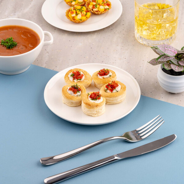 A white round plate with four small pastry appetizers, placed on a blue placemat alongside a fork and knife, with soup, a drink, and another plate of food in the background.