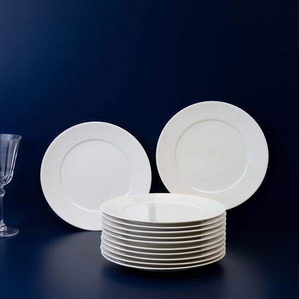A stack of plain white round plates is displayed in front of two upright plates against a dark blue background, with a clear glass visible on the left side.