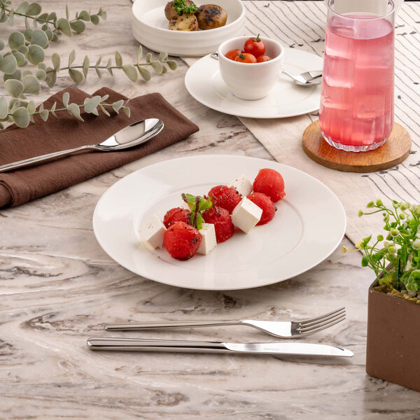 A round white plate with cubes of cheese and melon balls garnished with mint, set on a marble table with cutlery, a glass of pink beverage, and other small dishes nearby.