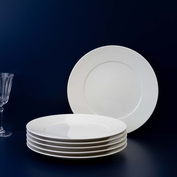 A stack of six plain white round plates is shown with one plate standing upright behind them and a clear glass on the left side, all set against a dark background.