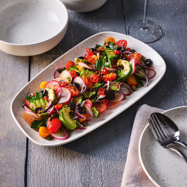 A rectangular white tray with speckled detailing, filled with a colorful salad of tomatoes, cucumbers, radishes, olives, red onions, and herbs, placed on a wooden table with plates, bowls, and cutlery nearby.