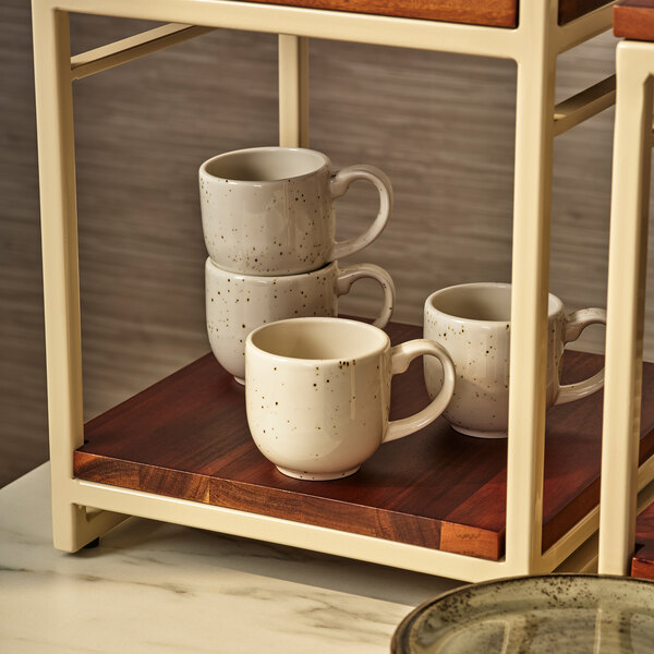 Four white ceramic mugs with a speckled pattern are displayed on a wooden shelf within a cream-colored metal frame.