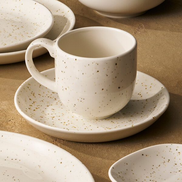 A white ceramic mug with brown speckles sits on a matching saucer, surrounded by similarly styled plates and bowls.