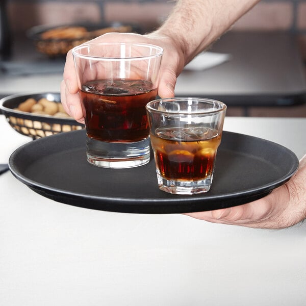 A person is holding a round black serving tray with two glasses of dark beverages containing ice.