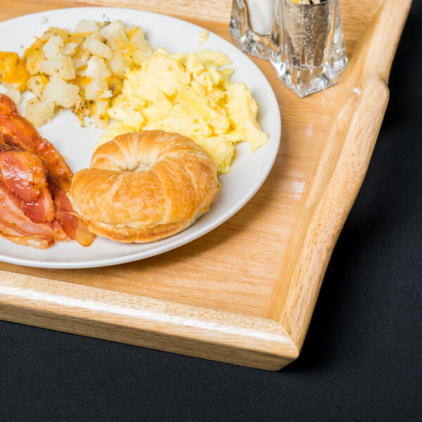 A wooden tray holding a white plate with scrambled eggs, diced potatoes, bacon, and a croissant, along with salt and pepper shakers.