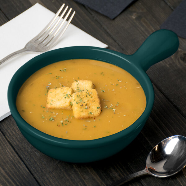 A Tablecraft hunter green cast aluminum soup bowl with croutons on top next to a spoon and fork.