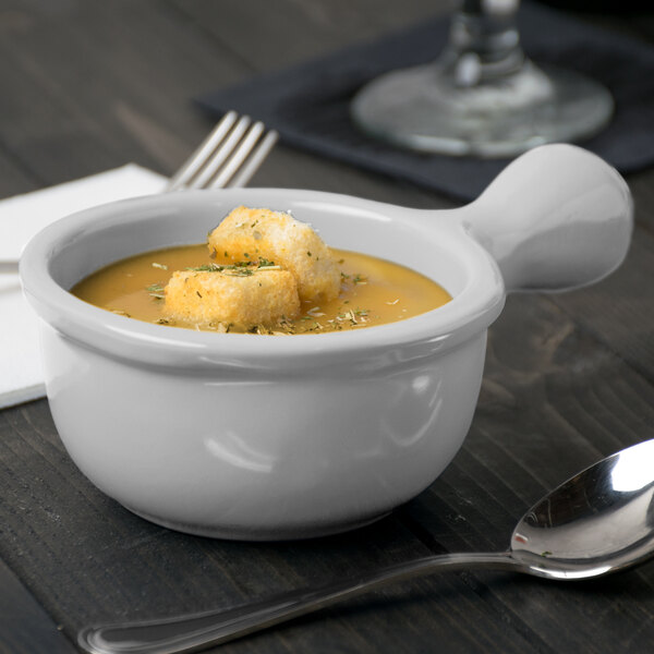 A Tablecraft natural cast aluminum soup bowl with a handle filled with soup and croutons with a spoon next to it.