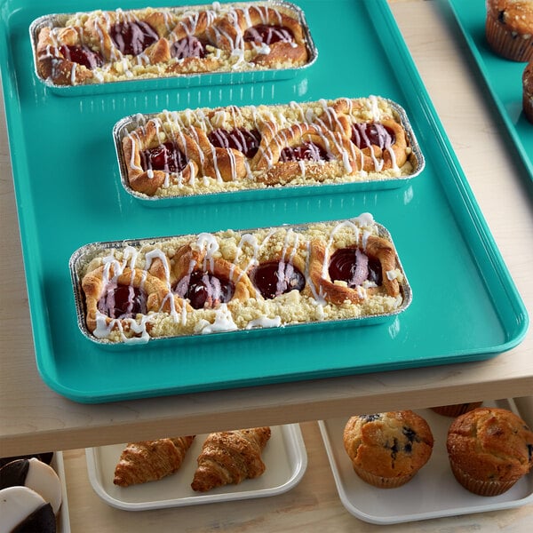 A green Cambro market tray of pastries and muffins on a bakery display shelf.