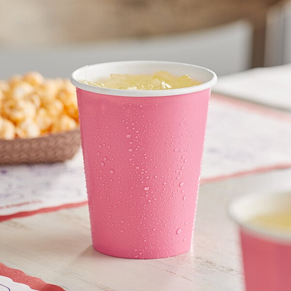 A Creative Converting candy pink paper cup with ice in it on a table with a bowl of popcorn.