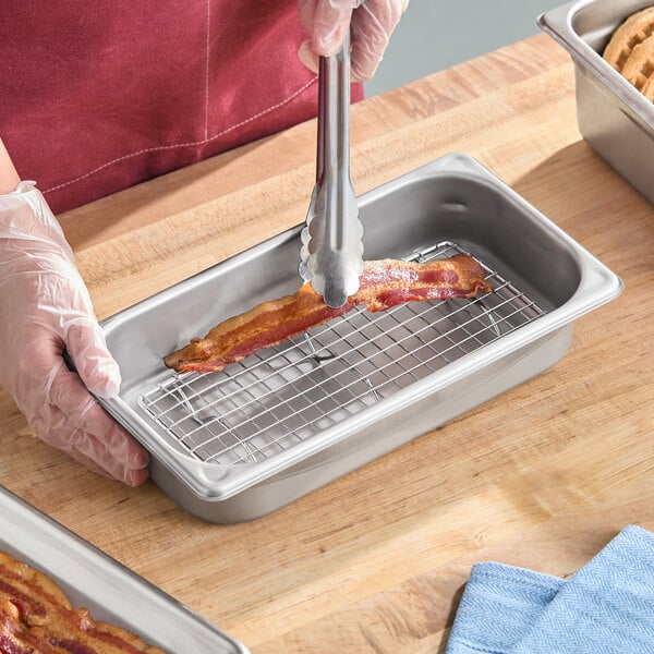 A small rectangular footed pan grate placed inside a steam table pan, shown holding a strip of bacon.