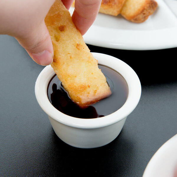 A person dipping a piece of bread into a white Tuxton ramekin of sauce.