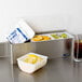 A San Jamar stainless steel condiment bar with a container of fruit and a drink on a counter.