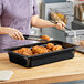A woman using tongs to place chicken in a Carlisle black plastic food pan on a counter.