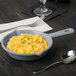 A bowl of macaroni and cheese in a gray Tablecraft fry pan on a table with a fork and spoon.