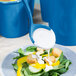 A Tablecraft sky blue cast aluminum long ladle pouring dressing onto a salad.