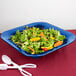 A Tablecraft cobalt blue cast aluminum square bowl filled with salad and oranges on a blue plate with spoons.