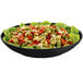 A Tablecraft black cast aluminum pasta bowl filled with pasta and lettuce on a table with lettuce and tortellini.