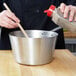 A hand pouring brown seasoning into a Vollrath stainless steel sauce pan.