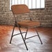 A National Public Seating brown metal folding chair with antique gold fabric padding set up in a room in front of a brick wall.