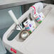 A gray Metro utility cart with three shelves and chrome posts holding a tray with a spoon, spatula, and measuring cup.