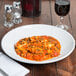 A white Tuxton porcelain bowl filled with pasta on a table with a glass of wine.