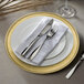 A white plastic charger plate with a gold rim and silverware on it.