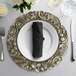 A silver Charge It by Jay Aristocrat charger plate on a table with a napkin and silverware.