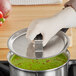 A person wearing gloves holding a Vollrath Tribute pan cover over a pot of green soup.