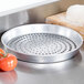 An American Metalcraft heavy weight aluminum pizza pan with perforations on the bottom, sitting on a table next to a tomato.