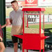 A man wearing a white shirt and beige apron uses a Carnival King red popcorn machine to fill a box with popcorn.