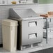 A stainless steel two-drawer mega top refrigerated sandwich prep table with a closed lid, positioned next to a beige trash bin in a commercial kitchen setting.